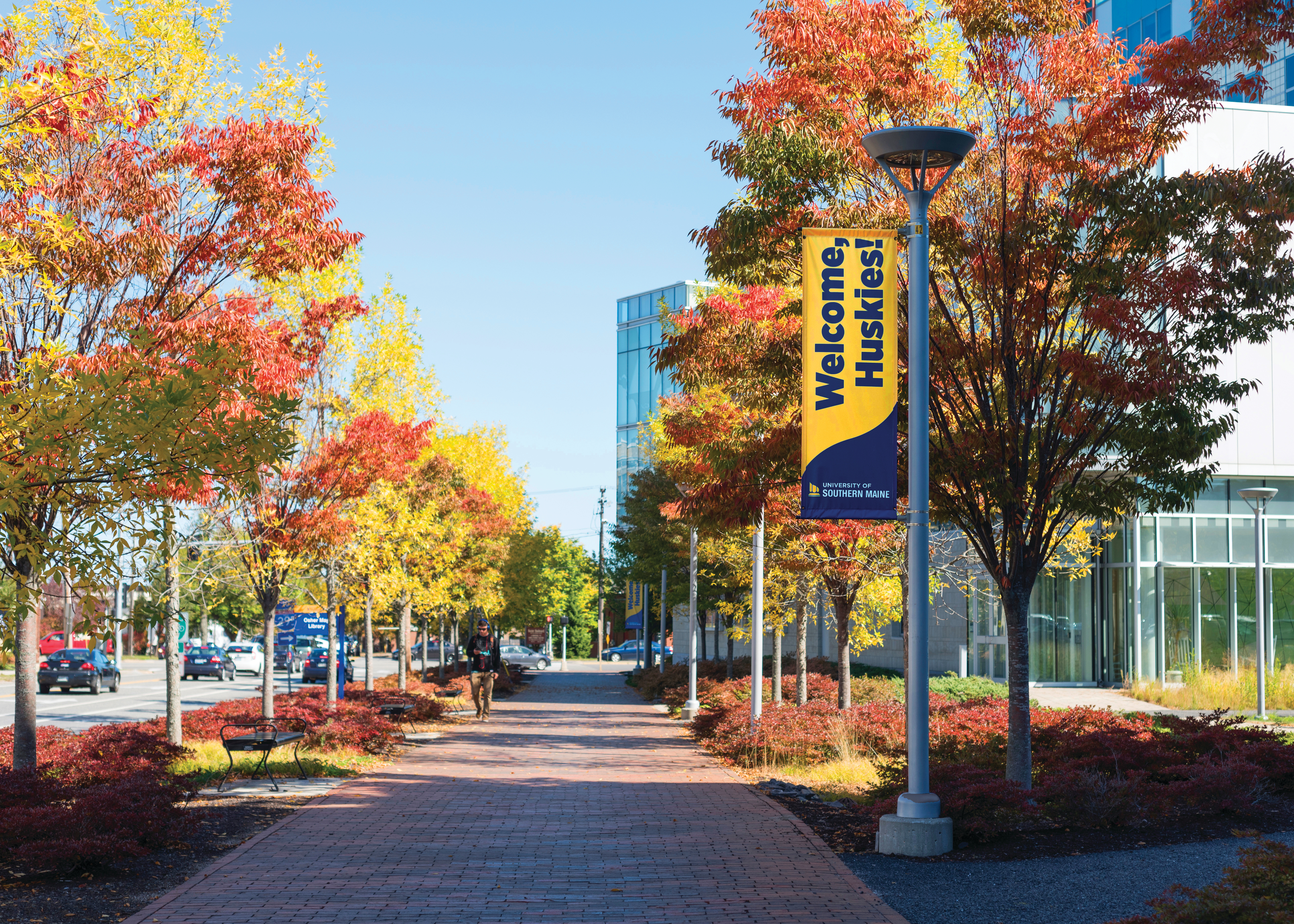 A photograph of the University of Southern Maine in the Fall. Looking down the sidewalk of Bedford Street in Portland towards the Glickman Library and Osher Map Library. A "Welcome Huskies!" banner hangs to the side.